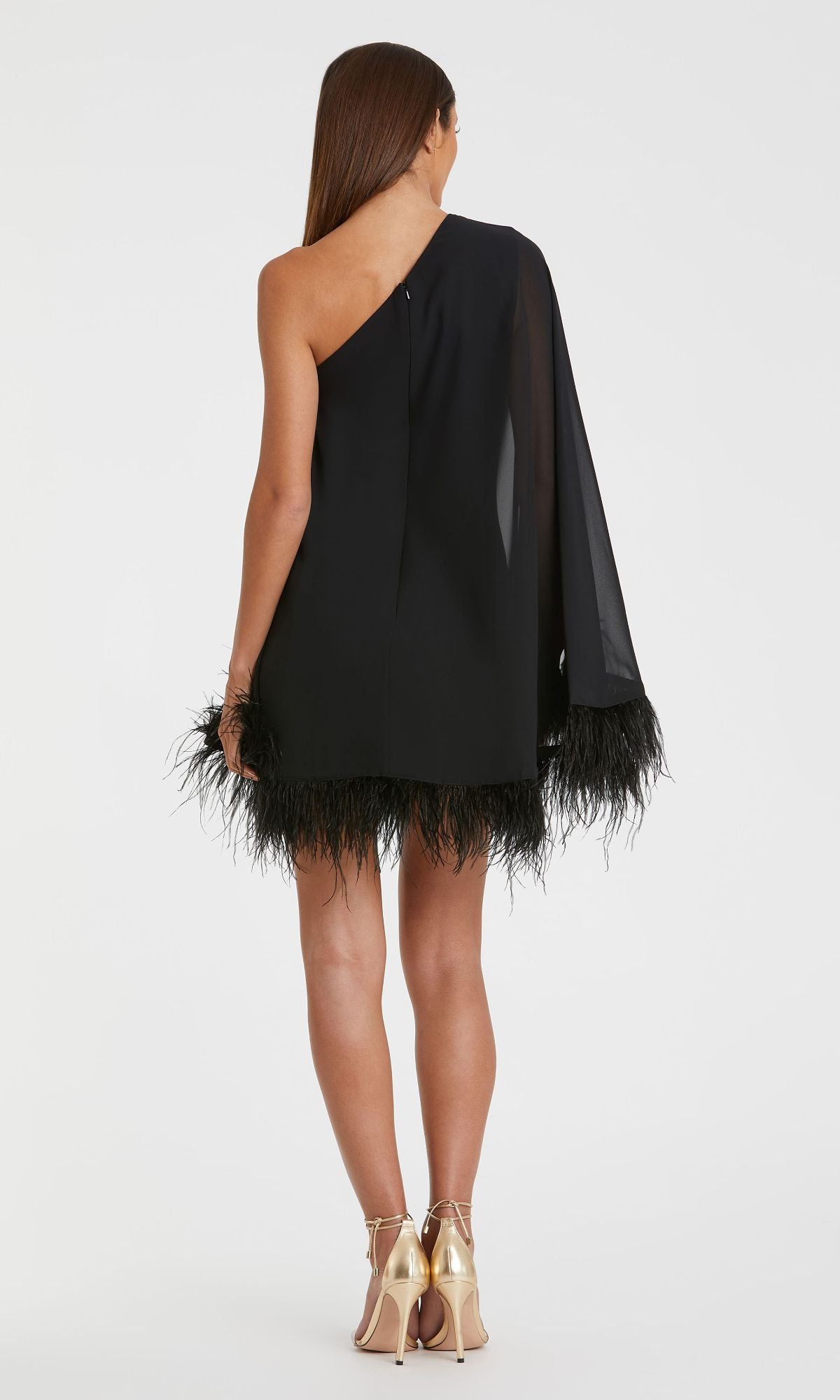 A person stands facing away, wearing the Mac Duggal 11623 short wedding guest dress—black, one-shoulder style with feather-trimmed hem and sleeve—paired with gold high-heeled sandals.